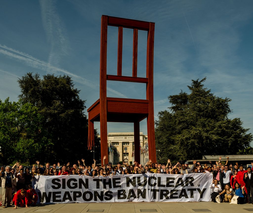 Fredsaktivister med en banderoll: "Sign the nuclear weapons ban treaty", vid den stora stolen med ett trasigt ben vid FN i Genève. 